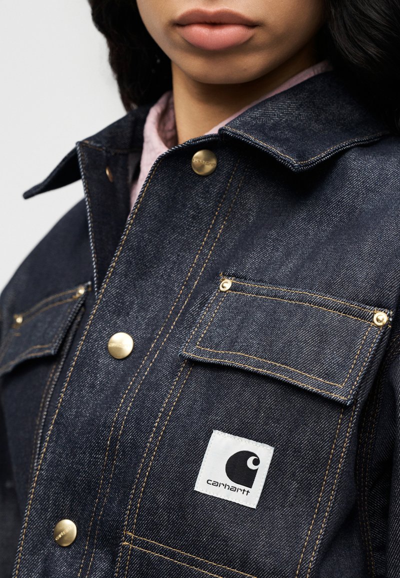 Veste en denim foncé avec ferrures dorées, deux poches avant et un écusson logo Carhartt. Les surpiqûres visibles ajoutent de la texture et des détails.