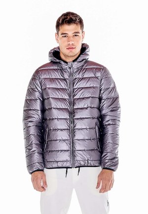 Un homme porte une doudoune grise brillante à capuche et un pantalon blanc sur un fond blanc uni.
