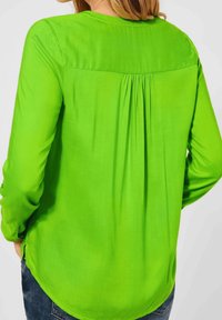 Grüne Bluse mit langen Ärmeln, Rückenfalte, runder Ausschnitt und glatter Textur. Kurviger Saum und taillierte Bündchen verbessern das Design.