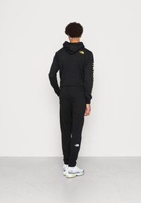 Černá mikina s kapucí a tepláky vyrobené z měkké tkaniny, na rukávu s logem North Face v žluté barvě. Doplňuje bílé a šedé sportovní boty.