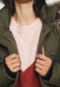 Olivgrön parka med pälsfodrad huva, bärs över en ljusrosa och röd färgblockströja. Tröjan har ribbad halsringning och mudd.