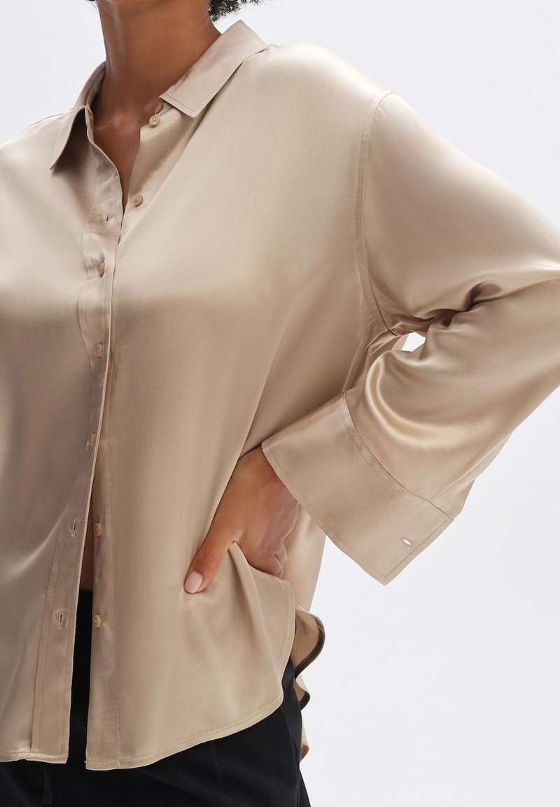 Femme portant un chemisier beige ample et brillant à boutons, manches longues, une main posée sur la hanche, sur un fond uni.