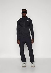 Uomo in piedi con giacca nera North Face, pantaloni neri, occhiali da sole neri oversize e scarpe da trekking bianche, su sfondo neutro.