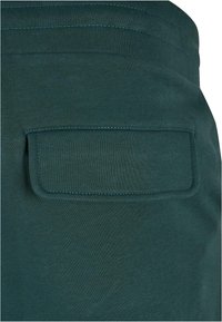 Țesătură verde închis cu o textură netedă prezintă un buzunar dreptunghiular aplicat. Accentele cosute într-o nuanță mai deschisă sunt vizibile pe margini.