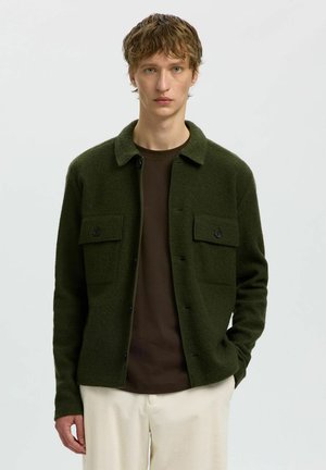 Jeune homme portant une veste-chemise vert foncé boutonnée, un t-shirt marron et un pantalon beige clair, debout devant un fond clair uni.