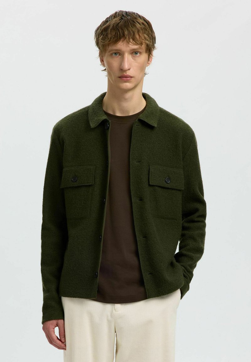 Jeune homme portant une veste-chemise vert foncé boutonnée, un t-shirt marron et un pantalon beige clair, debout devant un fond clair uni.