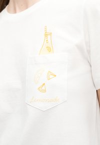 Λευκό μπλουζάκι με τσέπη στο μπροστινό μέρος που φέρει κίτρινες απεικονίσεις φέτες λεμονιού, ένα μπουκάλι λεμονάδας με καλαμάκι και τη λέξη «Lemonade».