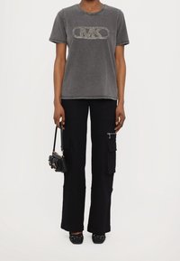 Camiseta de algodón gris con logo de strass, combinada con pantalones cargo negros con bolsillos y bailarinas negras con tachuelas. Sosteniendo un pequeño bolso negro.