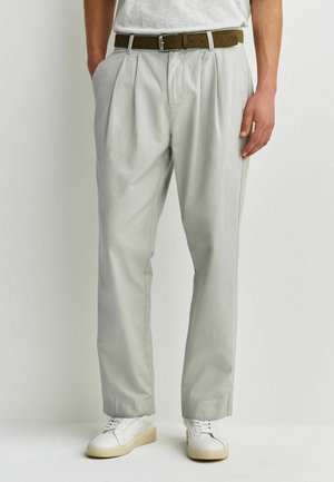 Mann trägt hellgraue, plissierte Hose mit braunem Gürtel und weißen Turnschuhen, steht vor einem einfarbigen Hintergrund.