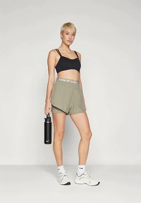 Čierny športový podprsenka, olivové zelené šortky s pásom "Nike Pro", čierna fľaša na vodu, biele ponožky a svetlé tenisky, stojace pred jednoduchým pozadím.