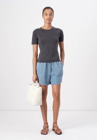 Black short-sleeve t-shirt, light blue denim shorts, and brown strappy sandals. Holds a white patterned bag. Minimalist design, soft textures.