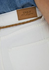 Bijele traperice od denima s kožnom etiketom na kojoj je ispisan tekst "ONLY Blue Denim", uz dodatak pletene smeđe kožne remenice. Prikaz u krupnom planu.