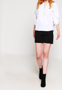 Frau in einer weißen, langärmeligen Schnürbluse, schwarzen Shorts und schwarzen Knöchelstiefeln, die vor einem schlichten hellen Hintergrund läuft.