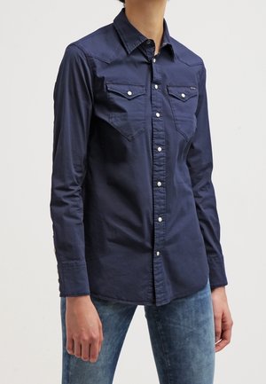 Person trägt ein eng anliegendes marineblaues Hemd mit Brusttaschen und hellblaue Jeans vor einem schlichten Hintergrund.