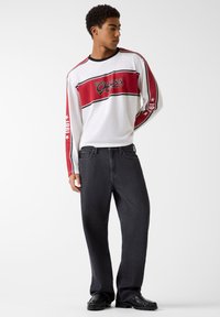 Biała siateczkowa koszula z długim rękawem z czerwonymi i czarnymi akcentami, z logo "Guess", w zestawieniu z luźnymi czarnymi jeansami i czarnymi butami.