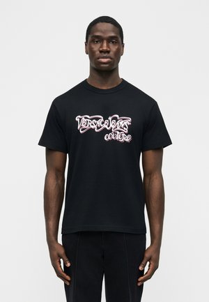 Bărbat tânăr stând cu fața înainte, purtând un tricou negru cu text stilizat alb și roz pe care scrie „Versace Jeans Couture.”