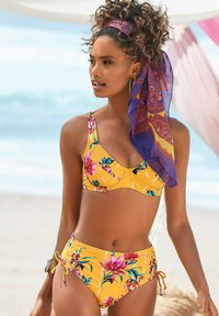 Haut de bikini floral jaune et bas taille haute assortis avec cordon de serrage. Écharpe en tissu transparent à motifs violets dans les cheveux, boucles d'oreilles créoles en or.