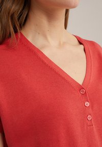 Czerwony sweter z dekoltem w serek, ozdobiony czterema jasnymi guzikami przy plisie. Gładka struktura z delikatnym detalem prążkowania.