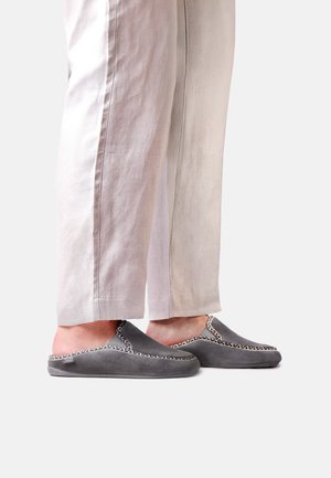 Personne portant un pantalon beige clair et des chaussons gris à enfiler avec une bordure à motifs, debout sur un fond clair uni.