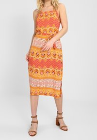 Šaty midi bez ramínek v oranžové barvě s květinovými a geometrickými vzory; stažený pas s zavazováním, vyrobené z lehkého materiálu, doplněné hnědými sandály na podpatku.