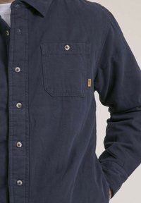 Chemise boutonnée bleu marine avec une poche poitrine unique, texture subtile et détails de boutons contrastants. Présente une coupe décontractée et des manches longues.