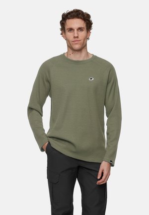 Mann in einem olivgrünen Waffelstrick-Langarmshirt mit kleinem Bärenlogo und schwarzen Arbeitshosen, steht mit einer Hand in der Tasche.
