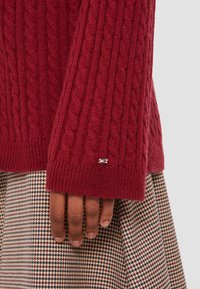 Main reposant à côté de la manche d'un pull en maille torsadée bordeaux avec un petit logo métallique, au-dessus d'une jupe ou d'un pantalon à motif pied-de-poule.