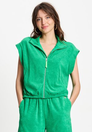 Jonge vrouw met bruin haar en creolen, die een groene mouwloze top met ritssluiting en bijpassende broek draagt, met haar handen in de zakken.