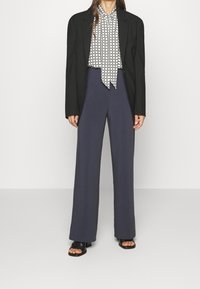 Blazer nero sopra una blusa bianca a fantasia con cravatta, abbinato a pantaloni navy a gamba larga e sandali neri con tacco.