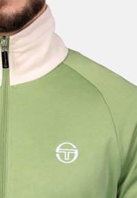 Grüne zip-up Jacke mit einem cremefarbenen Kragen, aus strukturiertem Stoff. Mit einem weißen aufgestickten Logo auf der Brust.