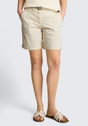 Persoon draagt beige shorts met opgerolde zoom, gevlochten riem en beige slippers met open tenen, staand met handen in de zakken.