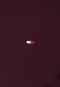 Bordeaux t-shirt met een geribbelde textuur, voorzien van een klein geborduurd logo in blauw, wit en rood, aan de linkerkant.