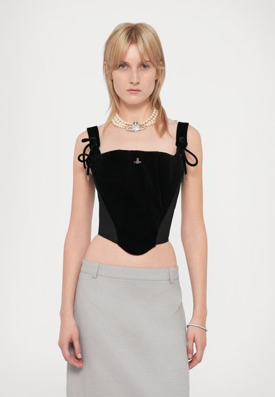 Blonde Frau trägt ein schwarzes Samt-Corsage-Top mit Schleifen-Trägern, eine Perlenkette mit Anhänger, einen grauen Rock und ein Armband vor weißem Hintergrund.
