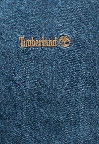 Timberland logo embroidered in brown thread on blue denim fabric.