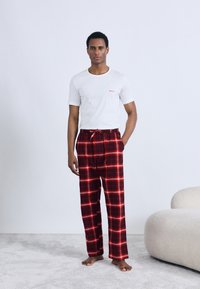 Λευκό βαμβακερό T-shirt με κόκκινο λογότυπο, συνδυασμένο με κόκκινο και μαύρο καρό παντελόνι πιτζάμα με ελαστική μέση και κορδόνι.