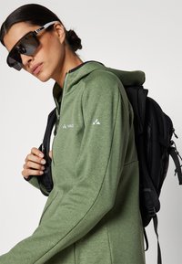 Sweatshirt verde com fecho, com um acabamento texturizado, apresentando uma mochila preta e óculos de sol oversized pretos com detalhes brancos.