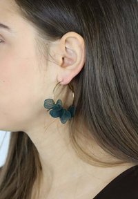 Złote kolczyki kółka z ciemnozielonymi akcentami kwiatowymi o teksturowanym wzorze, które komponują się z neutralnym tłem i długimi włosami.