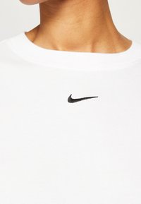 Vit bomulls-T-shirt med en svart broderad Nike-logotyp på vänster bröst, med klassisk rund halsringning och mjuk textur.