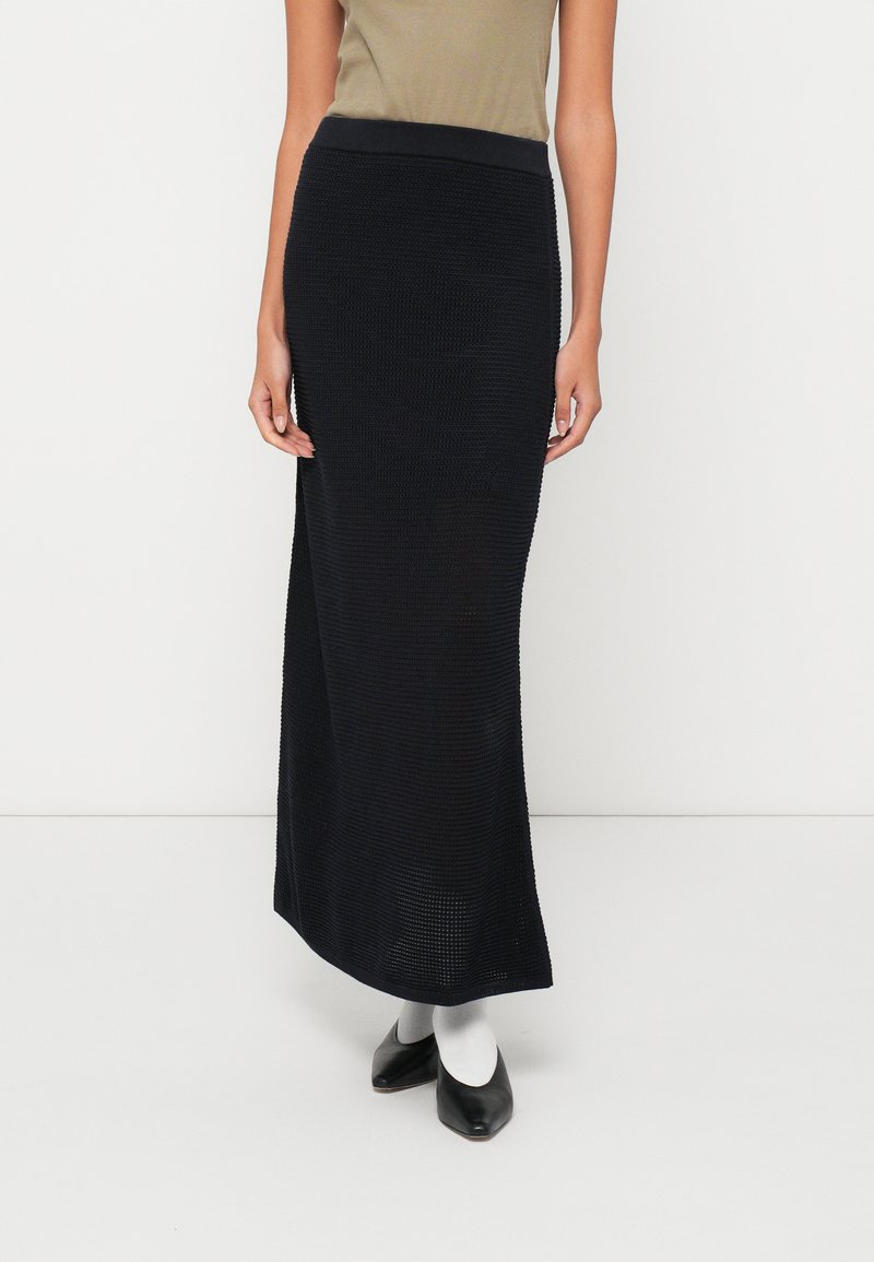 Fekete kötött maxi szoknya textúrált mintával, jól illeszkedő derékrésszel és egyenes szabással. Fekete hegyű cipővel és fehér zoknival párosítva.