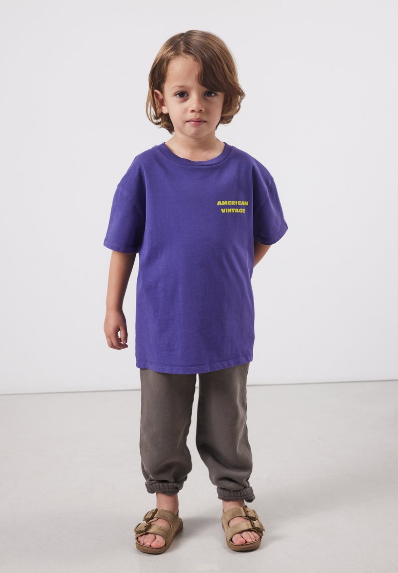 Jeune enfant aux cheveux mi-longs portant un t-shirt violet « American Vintage », un pantalon gris et des sandales marron, debout sur un sol blanc.