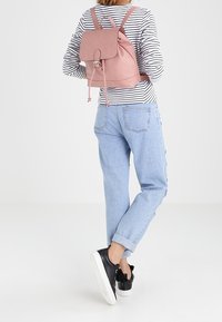 Person som bär ljusblå jeans, svarta sneakers, randig långärmad tröja och en rosa läderryggsäck, vänd bort från kameran på en enkel bakgrund.