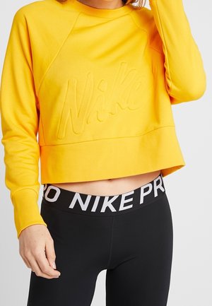 Vrouw draagt gele cropped Nike-sweatshirt en zwarte Nike Pro leggings, waarbij haar buik en hand dichtbij haar taille zichtbaar zijn.