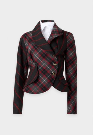 Femme portant un blazer à carreaux rouge et noir, une jupe noire brillante au genou, des talons noirs, tenant un petit sac à main noir devant un fond uni.