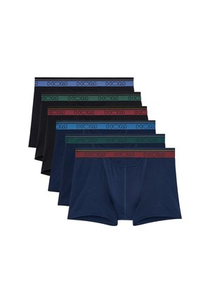 Mand stående barfodet på hvid baggrund iført sorte boxershorts med en mærket taljebånd, der kigger mod højre.