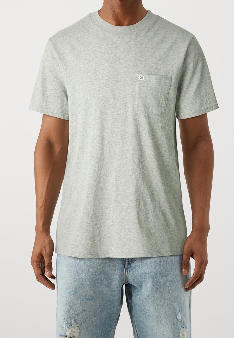 Homme portant un t-shirt gris clair à manches courtes avec une poche poitrine et un jean en denim bleu clair usé, debout devant un fond uni.