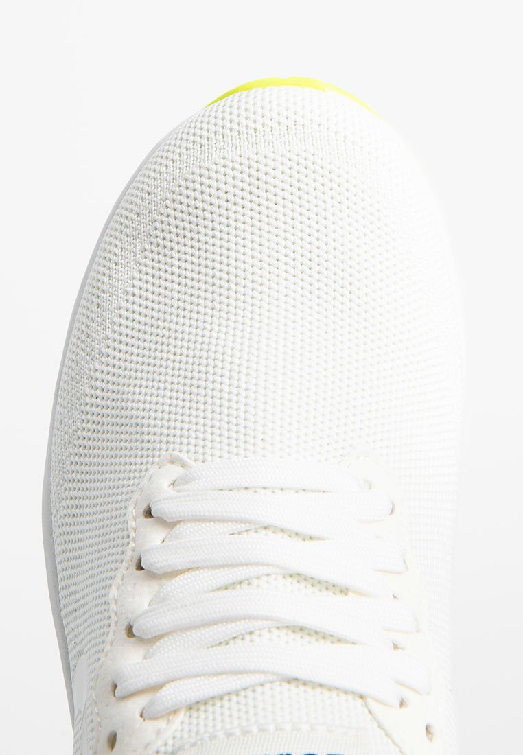 Zapatilla deportiva blanca con una parte superior de malla texturizada, con cordones blancos y un acento en el talón de un amarillo brillante. Diseño de puntera redondeada.