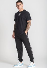 Svart bomullströja med en liten vit logotyp, tillsammans med svarta sweatpants med vita grafiska detaljer längs sidorna, och ljusblå sneakers.