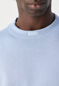 Svetlomodrá mikina s textúrovanou tkaninou, rebrovaným golierom a bielym vyšívaným logom "BOSS" na krku. Detailný pohľad.
