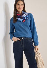 Femme portant un haut bleu à manches longues, une écharpe colorée et un jean foncé, tenant une veste en velours côtelé beige dans sa main gauche.