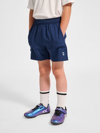 Marinblå idrottsshorts med elastisk midja och fickor, i kombination med iriserande lila sportskor och vita strumpor med svarta ränder.